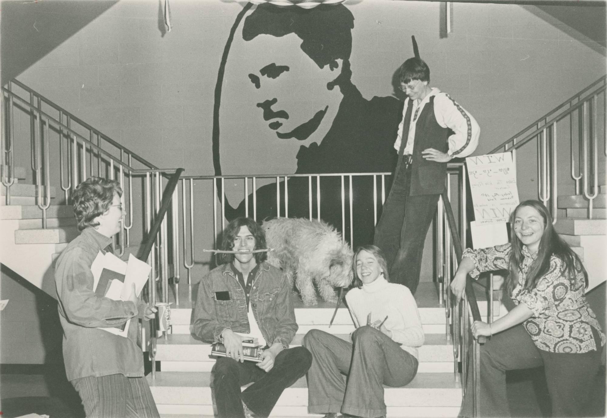 A black and white photo of students and faculty sitting and standing on the stairs in front of the William James mural in Lake Superior Hall.